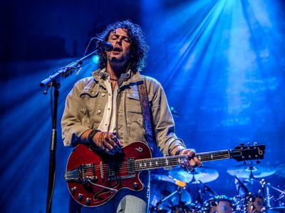 Lit by blue light a man with long brown curly hair sings into a microphone as he plays a red guitar