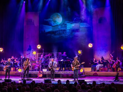 Four men playing guitars stand in a line across the front of the stage. Behind them are other musicians and a projection of a moon.