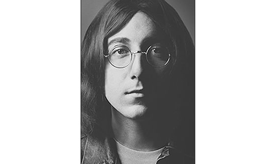 A black and white portrait of a person with long hair and round glasses, gazing seriously. The lighting casts a shadow on his face