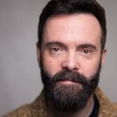 A headshot of James Mackenzie. He has dark hair with a beard and moustache and wears a black t-shirt under a beige cardigan