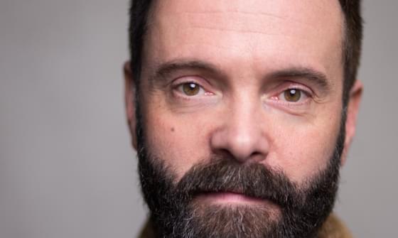 A headshot of James Mackenzie. He has dark hair with a beard and moustache and wears a black t-shirt under a beige cardigan