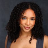 A black woman with long black curly hair smiling at the camera while wearing a black tank top