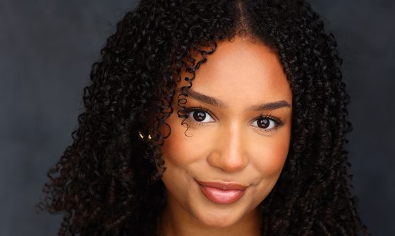 A black woman with long black curly hair smiling at the camera while wearing a black tank top