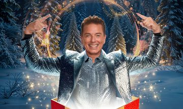 John Barrowman on a wintery forest background in a silver suit and shirt popping out of a red and yellow present smiling with his hands above his head pointing to all the golden glitter and magical looking circle around him.