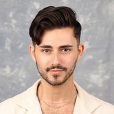 A young man with dark hair style into a quiff with stumbly facial hair, and a cream shirt that is open at the top exposing his chest hair and silver necklace, smiling softly at the camera