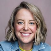 A white woman with a wavy blonde bob smiling at the camera with rosy cheeks, wearing a bright yellow top covered by a denim shirt