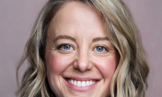 A white woman with a wavy blonde bob smiling at the camera with rosy cheeks, wearing a bright yellow top covered by a denim shirt
