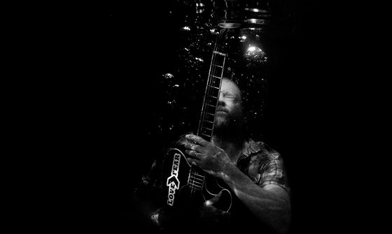 A black and white photo taken underwater of Nathan Bell holding his breath with his eyes closed and bubbles surrounding him while holding his guitar with a sticker saying 'Love>Fear" on it