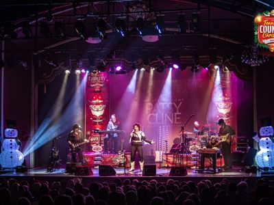 A vibrant Christmas-themed stage with musicians performing in cowboy attire, with decorative snowmen and festive lights