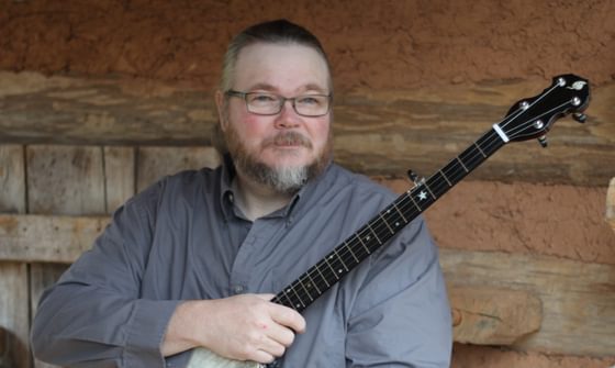 Riley stands against a wooden door holding a banjo. He has short hair, a moustache and beard, and wears a grey shirt