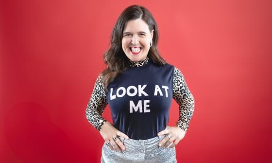 Rosie Jones stood smiling at the camera with her hands on her hips in a silver skirt and leopard print long sleeve top with a dark blue vest over the top with white text reading 'Look at me'