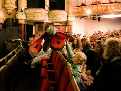 A photo inside of a grand theatre auditorium where a little girl is reaching up to touch a butterfly puppet
