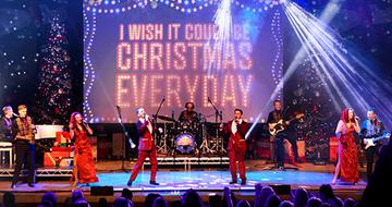 A festive stage performance with singers in red and gold, a backdrop reading "I WISH IT COULD BE CHRISTMAS EVERYDAY," Christmas trees, and bright lights