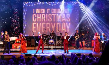 A festive stage performance with singers in red and gold, a backdrop reading "I WISH IT COULD BE CHRISTMAS EVERYDAY," Christmas trees, and bright lights