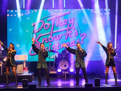 A lively stage performance featuring singers in coordinated outfits, with colourful lighting and a festive backdrop displaying "Do They Know It's Christmas?"