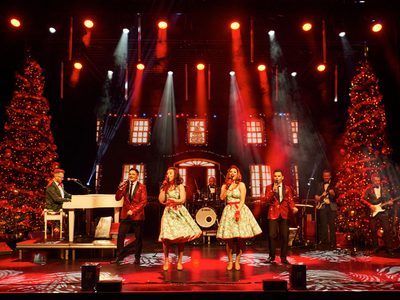 A festive stage performance with singers in vibrant outfits, surrounded by Christmas trees, twinkling lights, and a live band