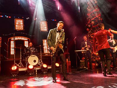 A saxophonist performs on stage in front of a festive backdrop with Christmas lights and a decorated tree