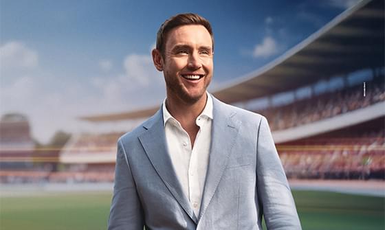 Stuart Broad smiling in a light blue suit, standing in a cricket ground with blurred stands and bright sky