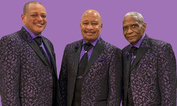 Three members of The Stylistics on a purple background in matching black suits with a purple pattern, purple shirts, and black ties, smiling at the camera