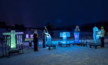 The Terje Insungset Ice Quartet stood outside at night in a snow covered field in large winter coats with their instruments made of ice around them lit by multicoloured lights