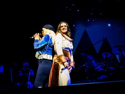 Two performers on stage dressed as Agnetha and Anni-Frid back to back with a band behind them in blue lights with a night sky projection