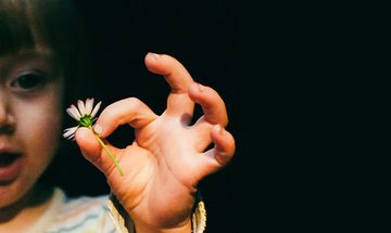 A little girl in a striped top holds a daisy between her index finger and thumb