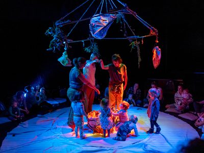 Lit by blue light five toddlers stand with the actors in the centre of the circle of adults and children