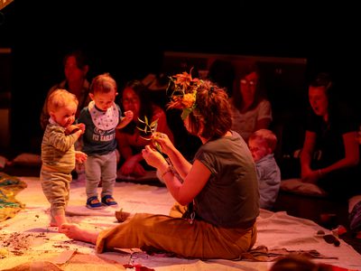 Two toddlers are interacting with a female actor who is sat on the floor in front of them