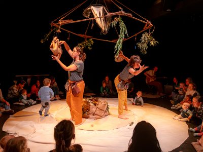 Two women in causal clothing perform in the centre of a circle of adults and children. One child can be seen running across the circle