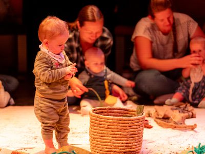 A toddler stands next to a woven basket and picks something out of it, behind the child there are parents and their children playing together