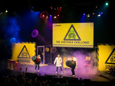 Three yellow warning triangles are projected on stage, reading The Airzooka Challange. Three scientists stand on stage using large air cannons to blast fog rings into the audience