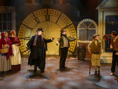 A stage dressed to be a Victorian street, with a shop front, and a front door, between them in the centre is a large fracturing clock. On stage are six actors, from left to right there is: two women in red dresses, an old man in a long black coat, colourful scarf, and top hat, a middle aged man in a beige shirt and brown top hat, a young boy using a crutch in a beige outfit with a scarf and flat cap, and a man in a brown jacket and green waist coat. Everyone is looking at the boy and smiling with him
