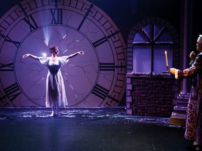 The stage is dark and lit in purple, with bright white light coming through the cracks in the large clock. In front of the clock is a white woman in a white dress posed like a ballerina. The old man in the night cap and robe is stood to the right holding a candle stick and looking shocked
