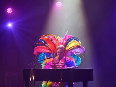 An Elton John impersonator performs at a piano on stage, dressed in a colourful suit decorated with feathers.