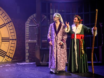 Two old white men on stage. One is in a white night cap and long purple robe, he's stood next to the other man who is in a long green robe, with white fur trim and a red belt, he is also wearing a holly crown and holding a large staff