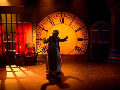 The stage is dark, only lit at the back with orange and the clock is illuminated orange. There is a silhouette of an old man in a night cap and long gown and robe moving across the stage