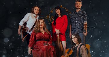 Five people all sat and stood on a dark background with snow falling, they all look very serious. There are three people stood, two white men both holding guitars and a white woman between holding a dulcimer, a long string instrument. There is one white woman sat on a chair holding a wooden star, and a white woman on the floor holding a violin