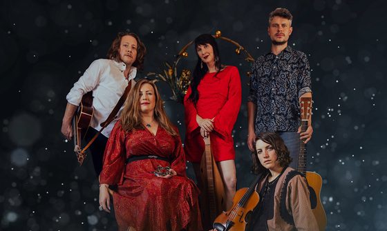 Five people all sat and stood on a dark background with snow falling, they all look very serious. There are three people stood, two white men both holding guitars and a white woman between holding a dulcimer, a long string instrument. There is one white woman sat on a chair holding a wooden star, and a white woman on the floor holding a violin