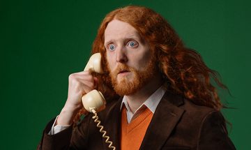 Alasdair Beckett-King, a pale white man with long curly ginger hair, a short ginger beard, and bright blue eyes. He's wearing a brown jacket with a white collared shirt and an orange jumper. He looks shocked, holding a corded phone to his ear