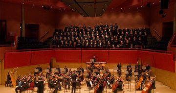 A choir performs behind an orchestra on stage, with a conductor leading the musicians and singers, all dressed in formal black attire.