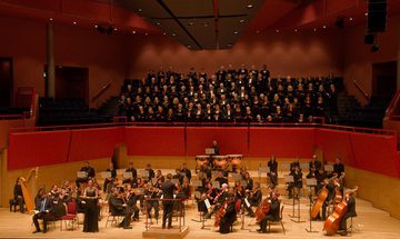 A choir performs behind an orchestra on stage, with a conductor leading the musicians and singers, all dressed in formal black attire.