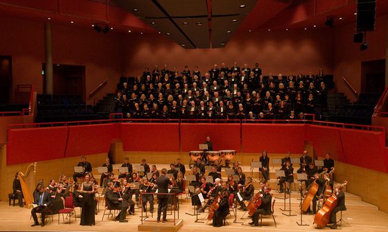 A choir performs behind an orchestra on stage, with a conductor leading the musicians and singers, all dressed in formal black attire.
