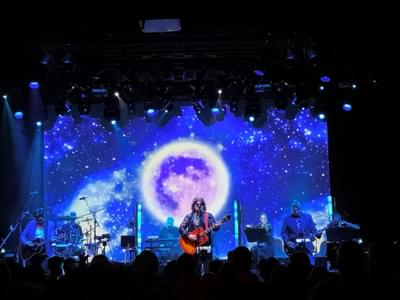 The ELO band performing on stage with a large projection of a moon on a galaxy sky behind them