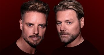 Keith Duffy and Brian McFadden stood together with neutral expressions in black t-shirts on a dark red background
