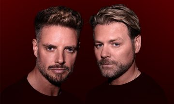 Keith Duffy and Brian McFadden stood together with neutral expressions in black t-shirts on a dark red background