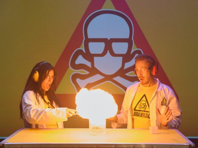 A male and a female scientist wear lab coats and hear defenders as they light an explosion before them. Behind them is a red warning triangle with a skull in it