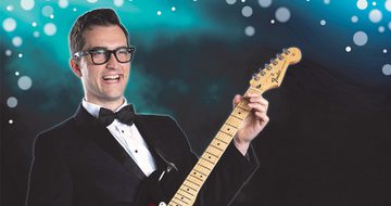 A man wearing glasses, a suit and a bow tie plays guitar against a blue, sparkly background.