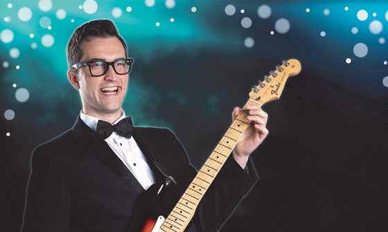 A man wearing glasses, a suit and a bow tie plays guitar against a blue, sparkly background.