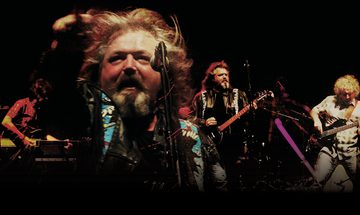 A dark image of three men on stage playing guitars. In the centre left is a massive image of a man with long hair and a beard with a microphone in front of him