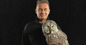 A close-up of Chris Packham who is wearing a black jumper. He is holding an owl that is staring forwards.